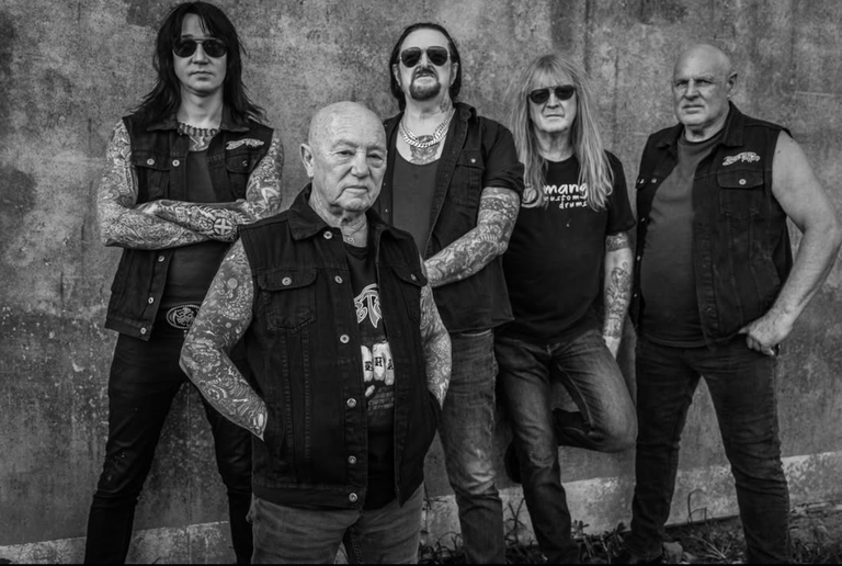 Black and white portrait of Australian rock band Rose Tattoo. The five heavily tattooed members pose in black denim vests against a concrete wall.